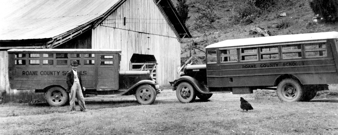 Historic photo of two buses with Roan County Schools written on them by a barn with a person to the side and a chicken in front of a bus.