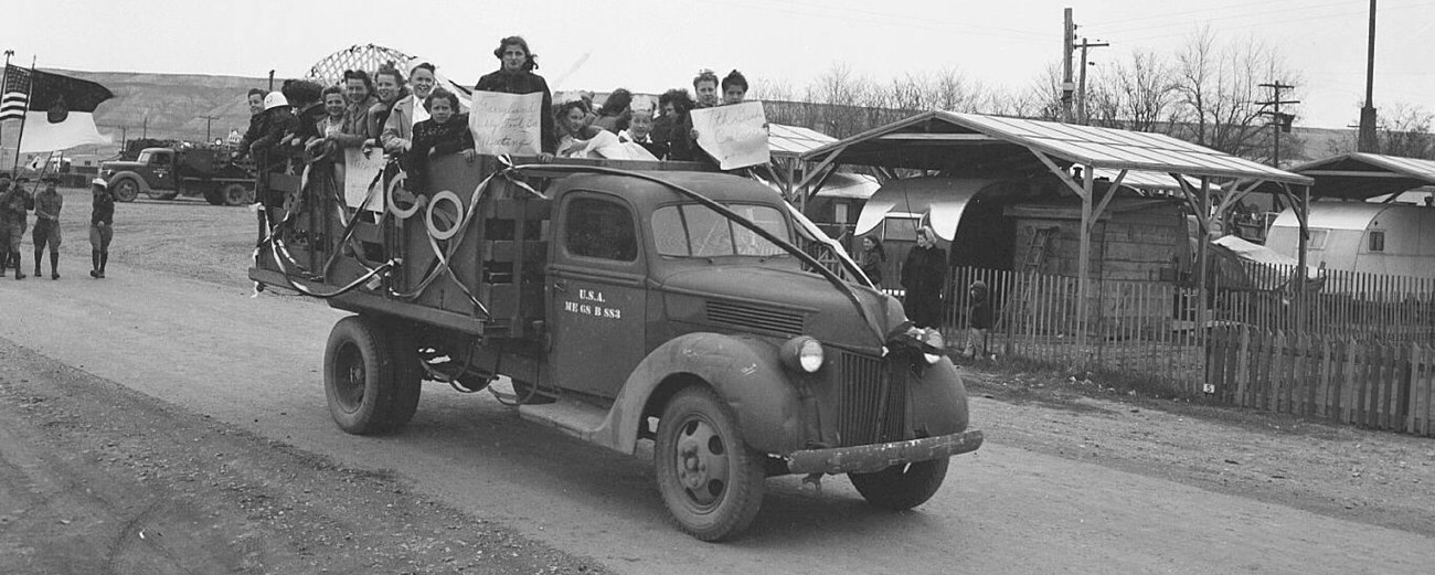 1940s truck with around 16 older kids standing in the back holding signs driving on a dirt road.
