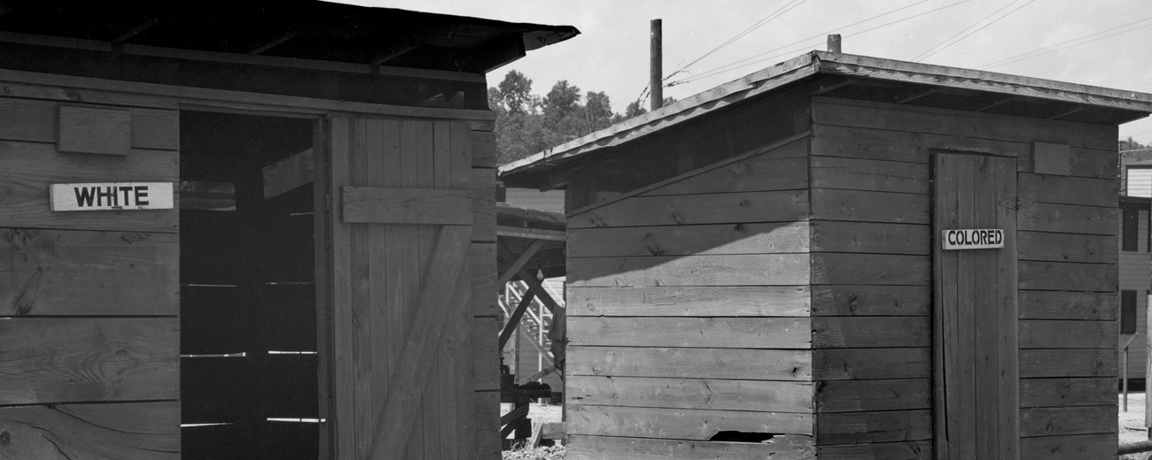 A black and white photo of two small wooden shed-like buildings side by side. One has a sign that reads “WHITE” while the other has a sign that reads “COLORED.”