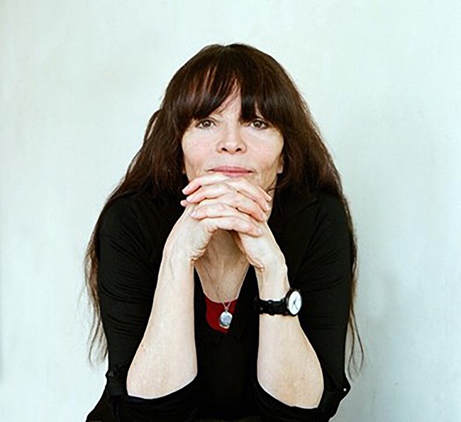 Trisha Pritikin A women with long dark hair is seated resting elbows on knees and hands interlaced in front of chin looks directly at the camera
