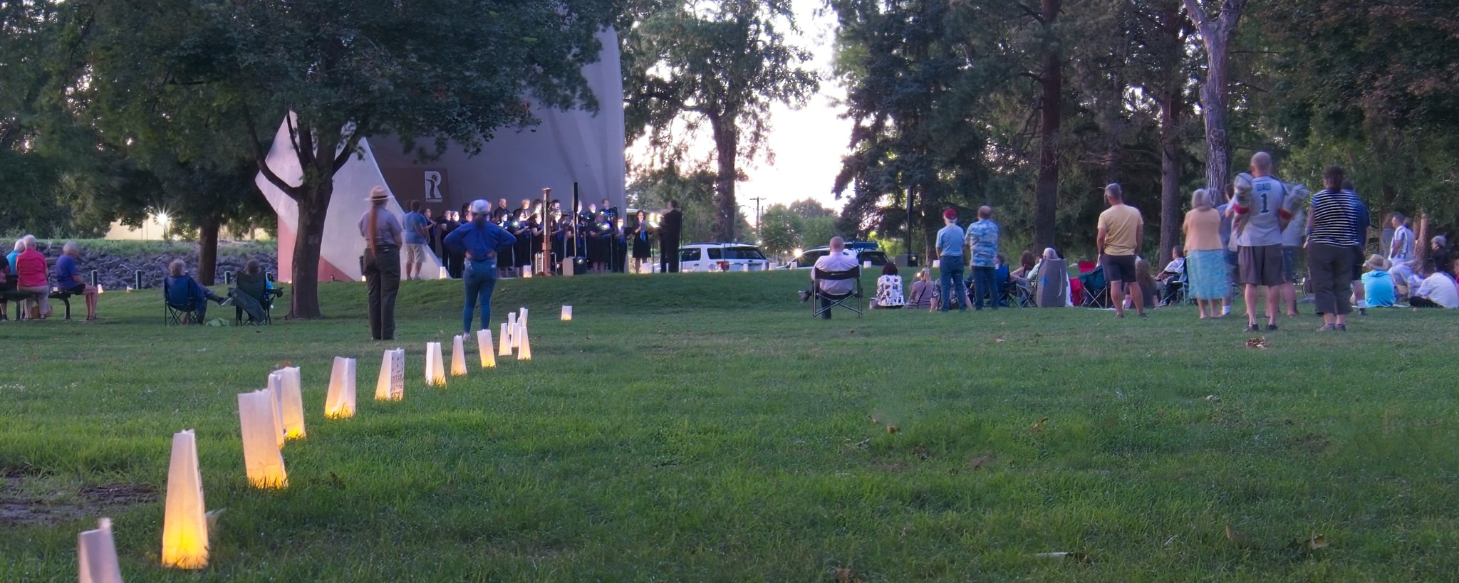 Illuminated paper bags line a grassy field, people gather at the far end in the fading daylight.