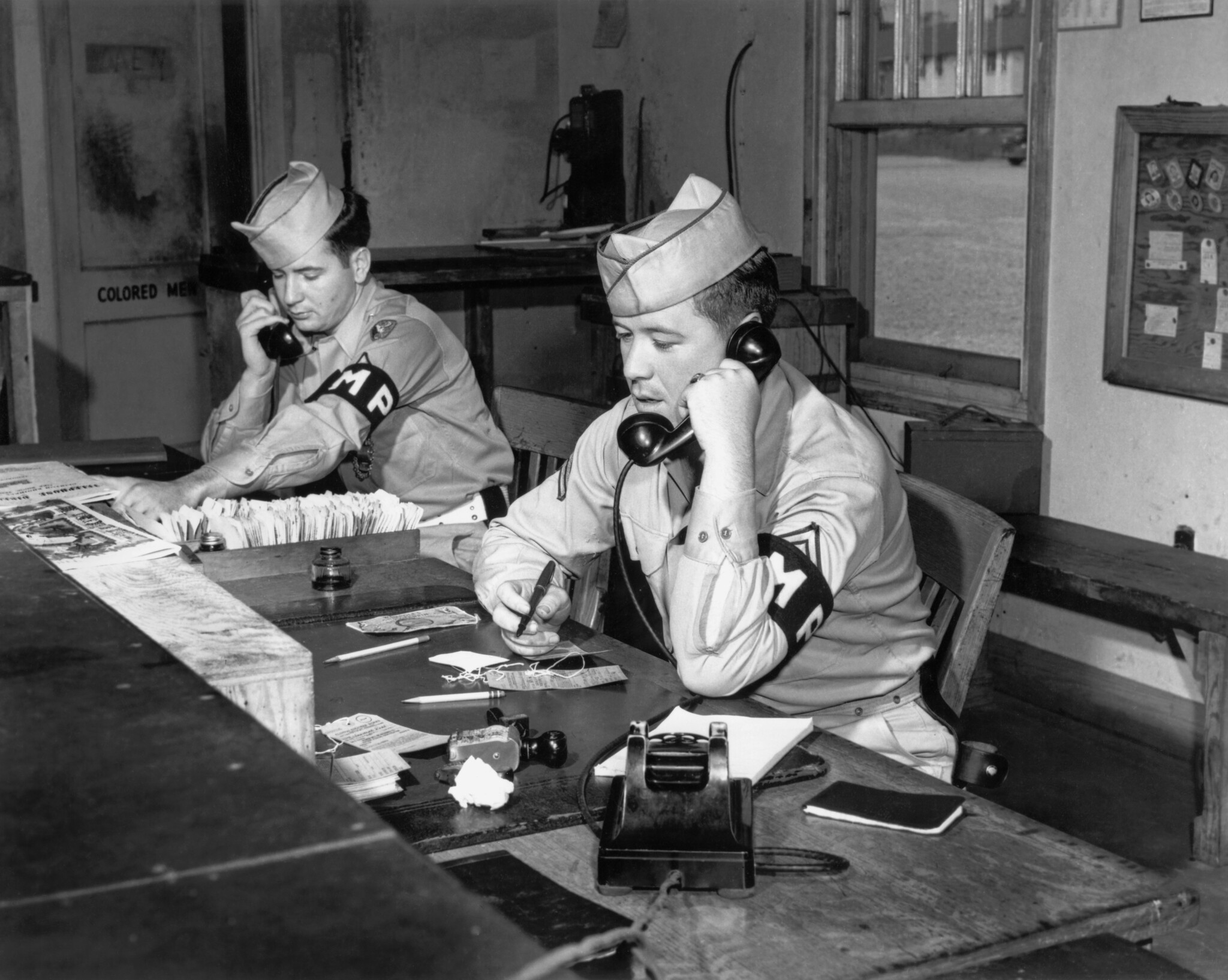 Two Military Police sit at a desk talking on phones in a guard house.