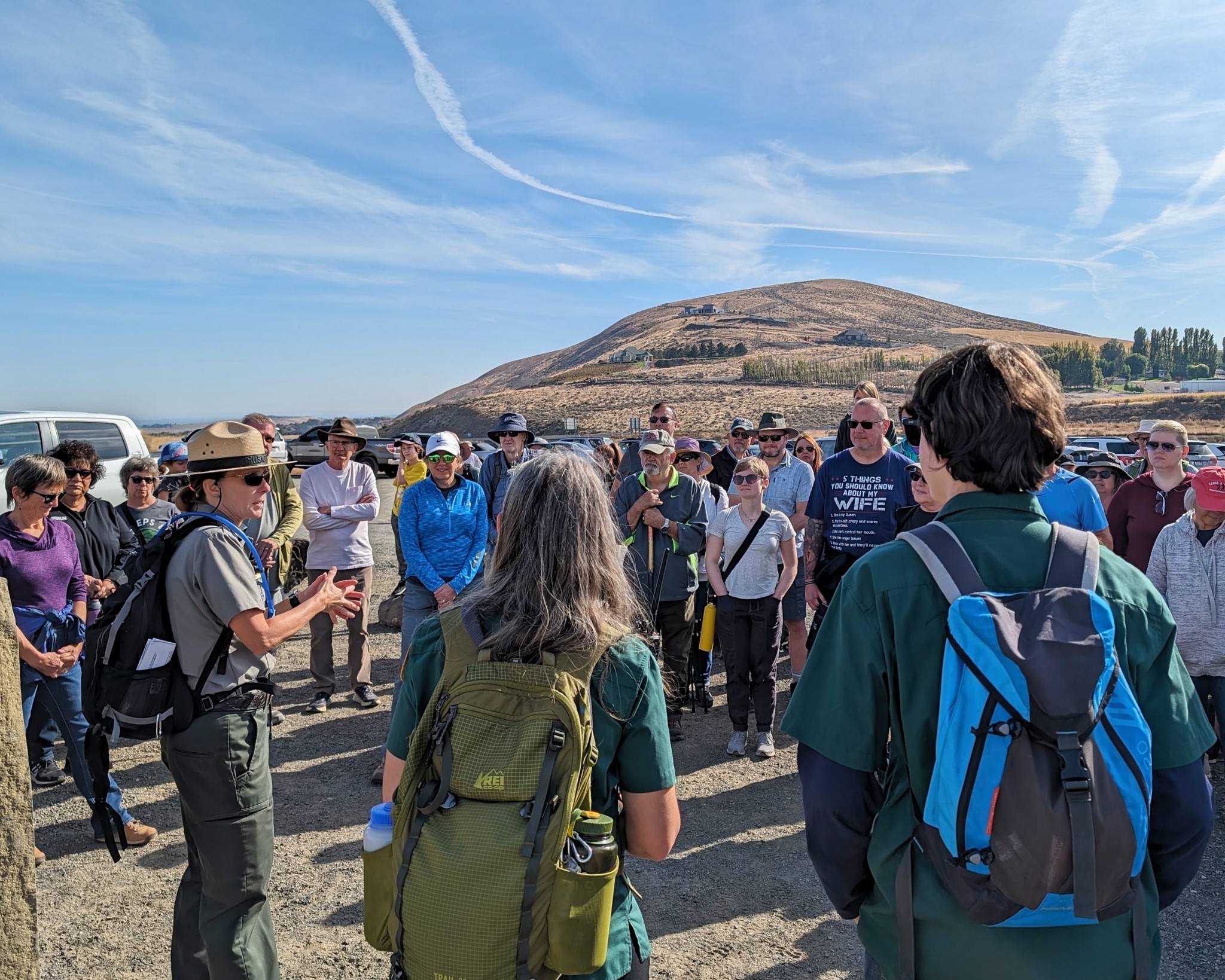 A woman wearing a campaign hat and green jacket and pants speakers to a group of more than 50 people. Blue sky and brown hills fill the background.