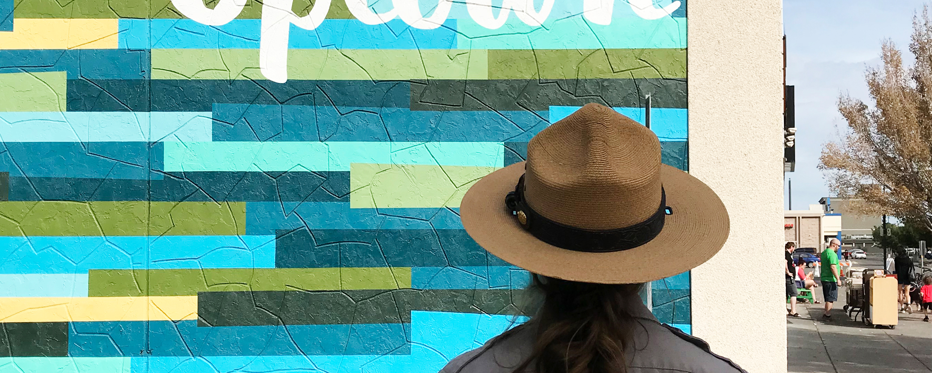 A color photo of a woman in a flat hat standing before a building with the word "Uptown” in white on green and blue block graphic background.