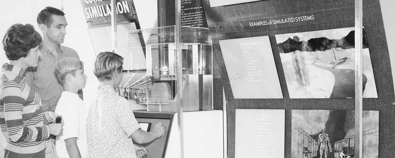 Black and white photo of nuclear family standing at a wall-mounted museum exhibit with a metallic box and wall panels with images and text, “Examples of Simulated Systems.”