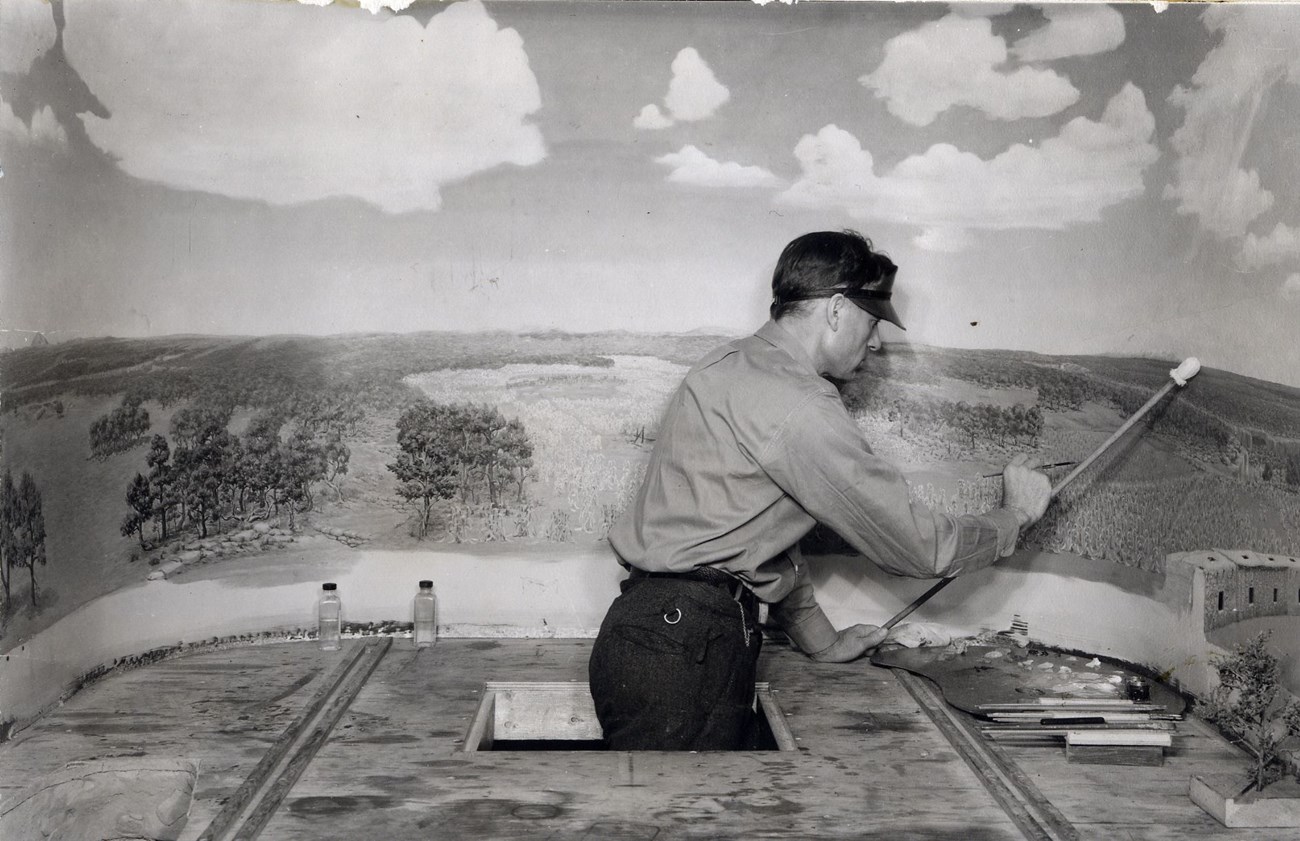 Historic photo of a man inside a diorama box. He is wearing a visor and finishing painting the sky on a scene of farms and forest