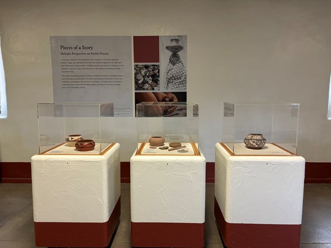 temp exhibit Pieces of A Story Three square display cases, dark red bottom, white middle third and  tall glass on top. These contain ancient and modern pottery. On the wall hangs a large exhibit about pottery