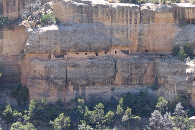 Inside an alcove within tall sandstone canyon walls is a partially built dwelling with walls, doors and windows of hand carved stones