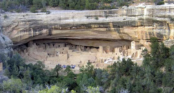 Cliff Palace alcove. a large rock overhang where people built square and round buildings out of stone blocks. A group of people stands in the center, showing the massive scale.