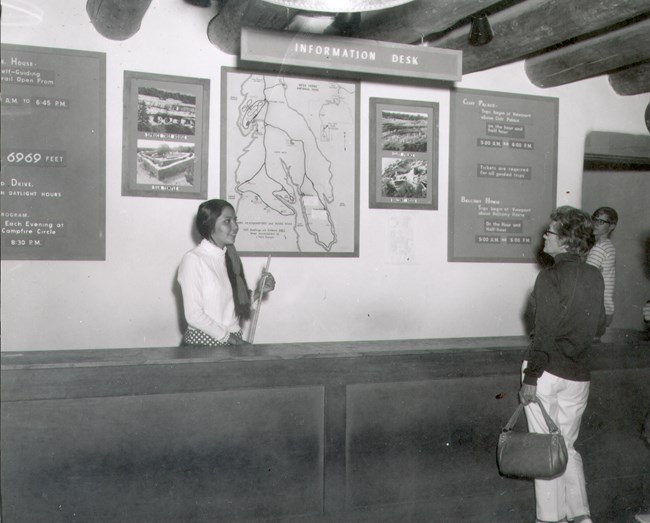 Historic photo of the museum information desk A young woman smiles at a park visitor from behind the museum information desk with maps and tour information posted on the wall