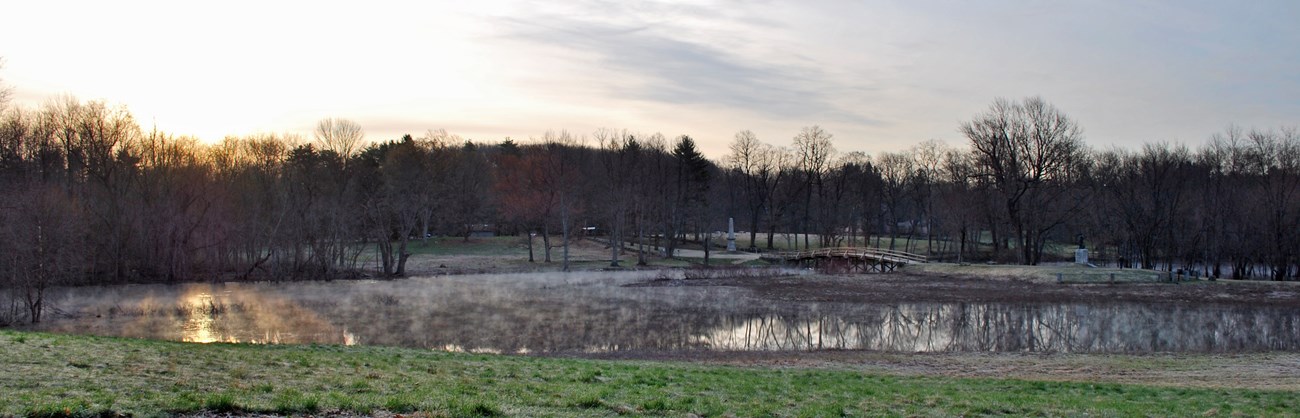 A scenic view of the Concord River illuminated by early morning sunlight. The wooden North Bridge is seen arching the river covered by a thin layer of fog. Rolling hills of green grass surround and towering trees are seen in the distance.