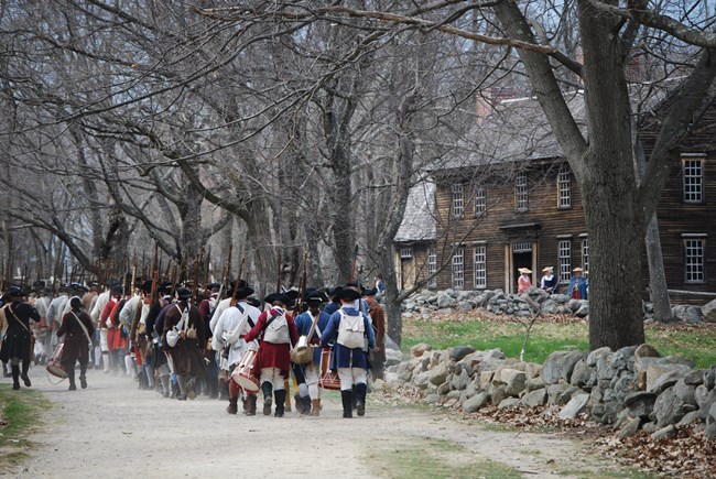 A long column of Colonial Militia with muskets and equipment mark down a dirt road flank with stonewalls on either side. The soldiers march past a wooden colonial house where women stand near the doorstep