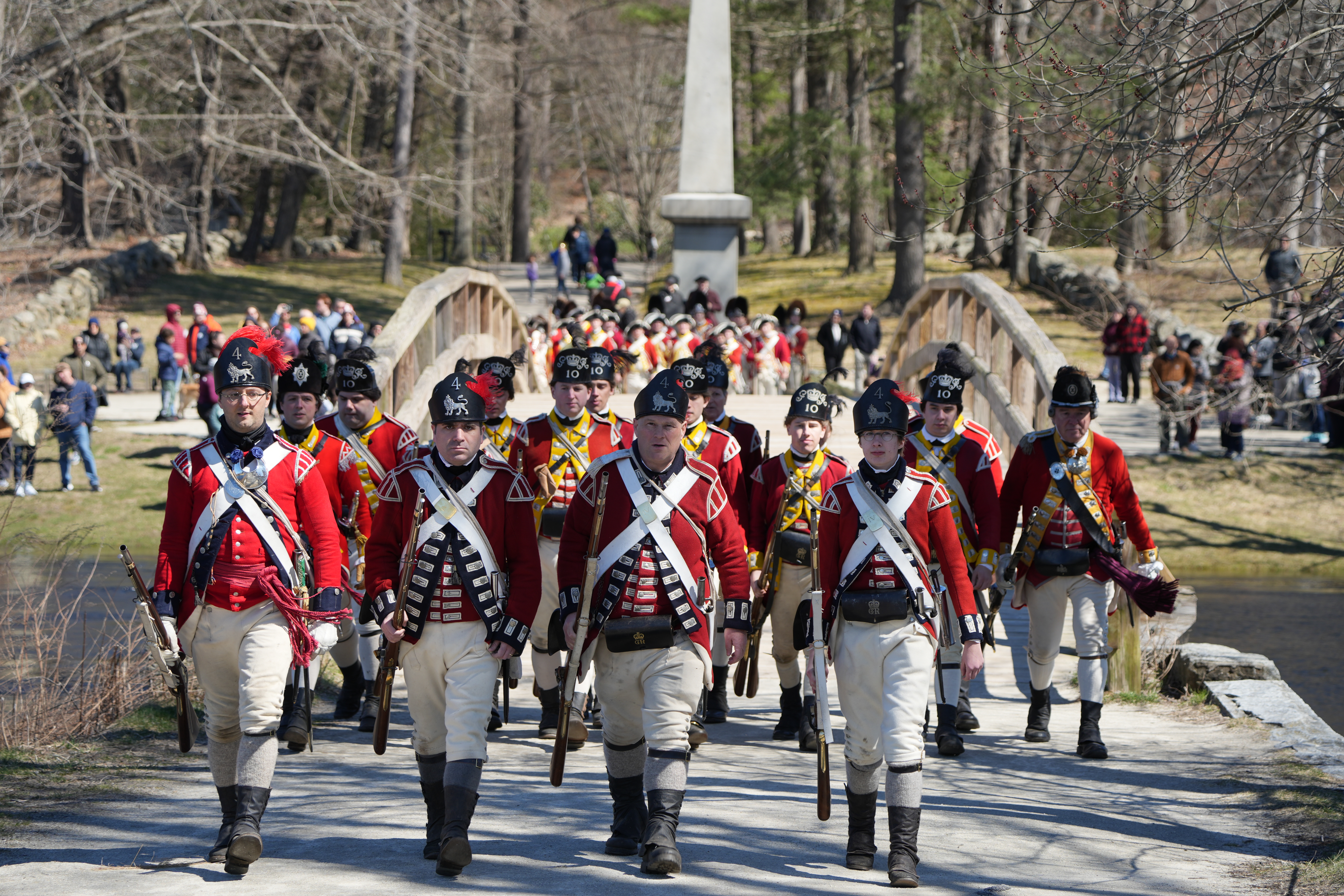 British redcoat soldiers march in line with muskets over a wooden bridge in Concord, Ma. Park visitors watch the soldiers from the distant riverbank in the background.