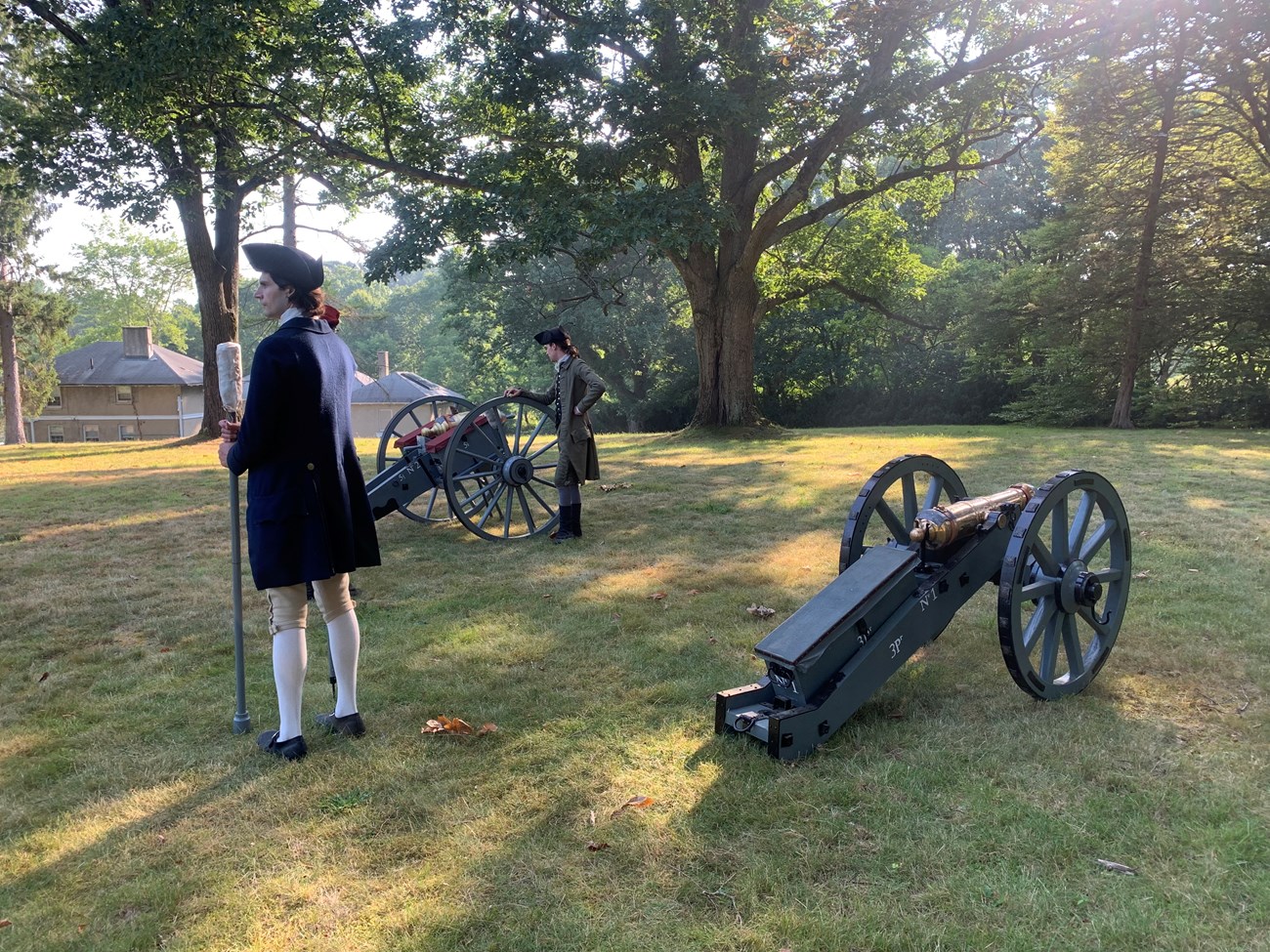 Two small cannon sit in a grassy field while two militia artillerist in 1775 clothing stand nearbye