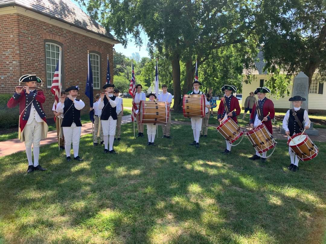 Group of young colonial musicians holding instruments standing in front of a brick building.