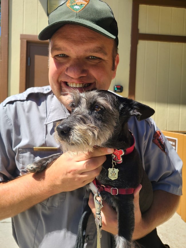 Man wearing park ranger uniform holding a small black dog.