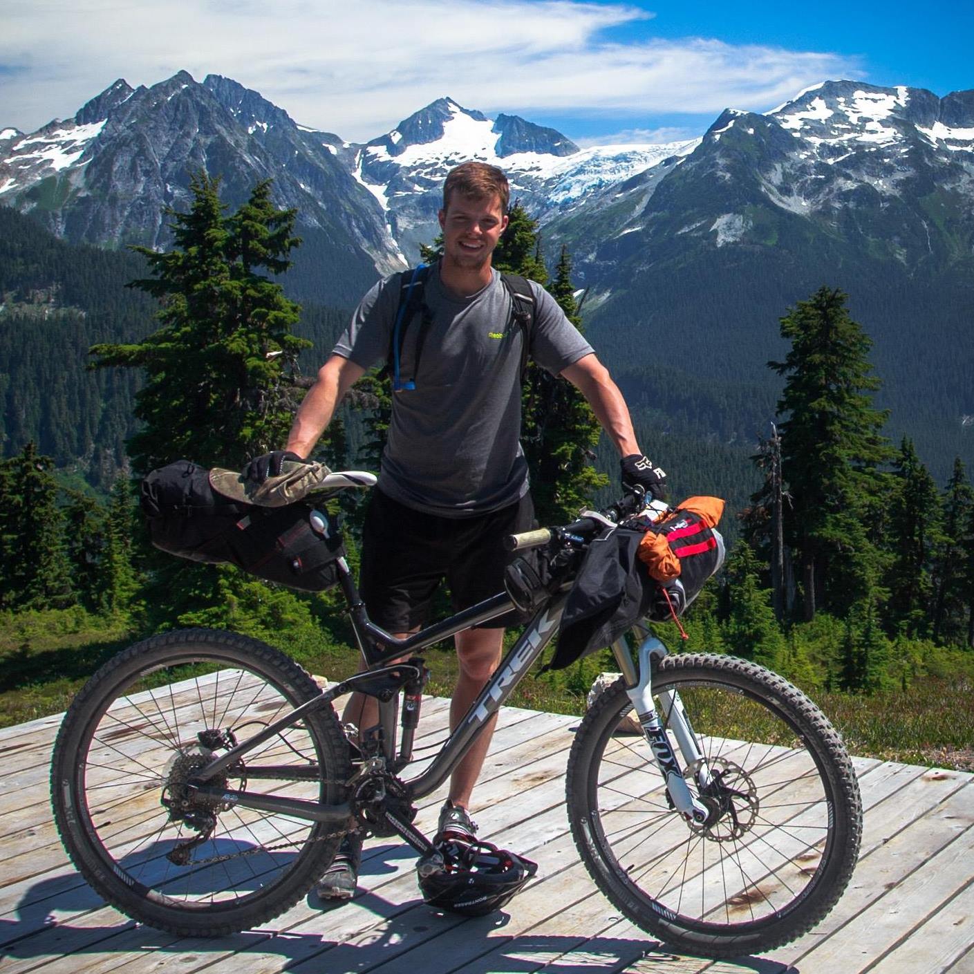 Matthew Bunker stands with his bicycle with snowcapped mountains in the background.
