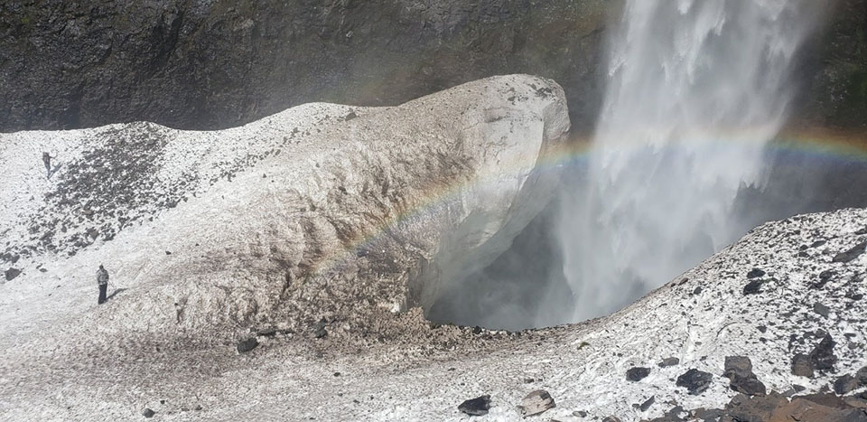 Two searchers on snow near a large hole that waterfall pours into with a rainbow in its mist.