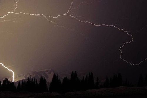 A lightning bolt zigzags across a dark sky above the slope of a mountain.