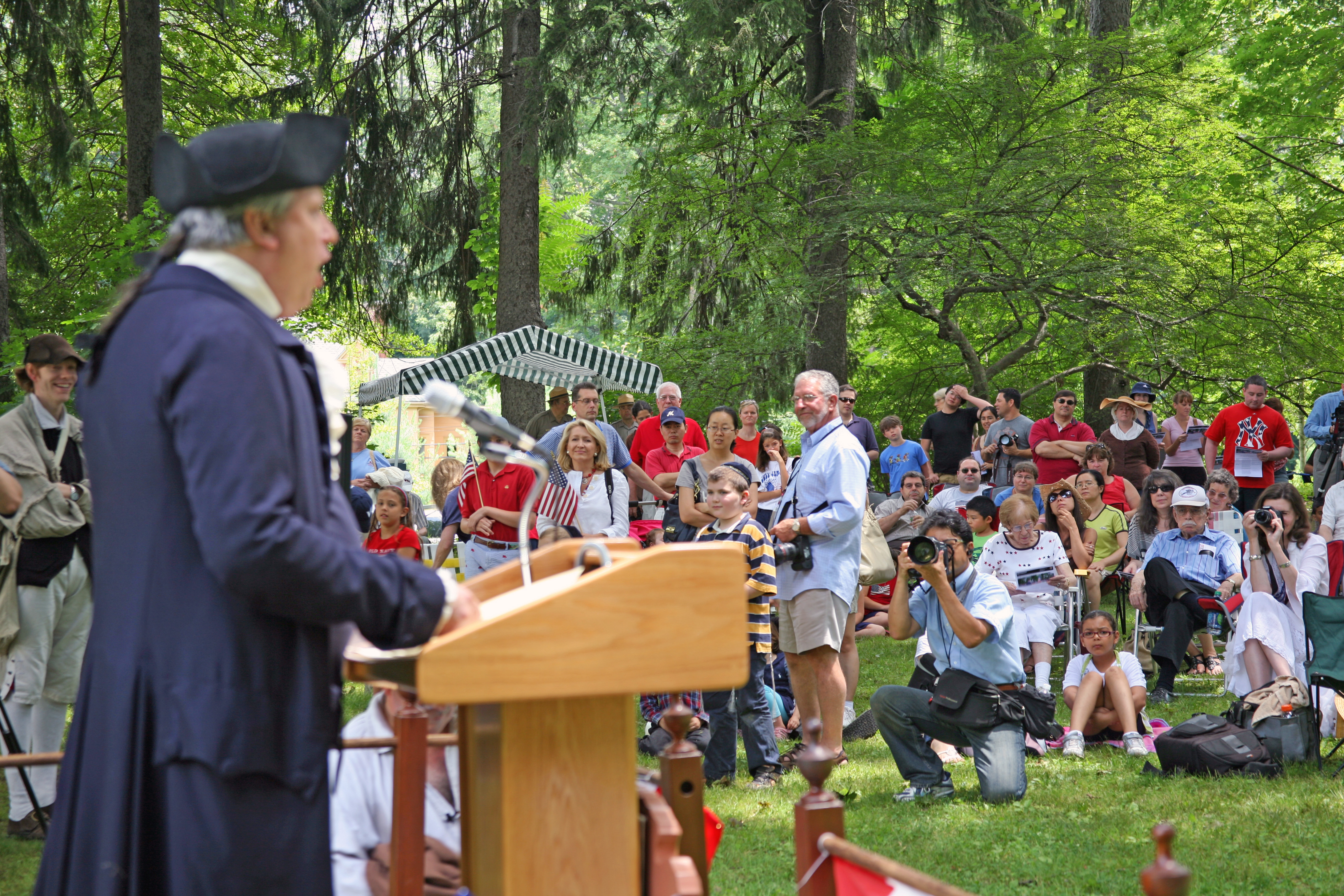 A speaker in colonial attire addresses a diverse crowd during an outdoor historical event. The audience includes children and adults, some holding American flags and taking photos, with a backdrop of trees and a canopy providing shade.