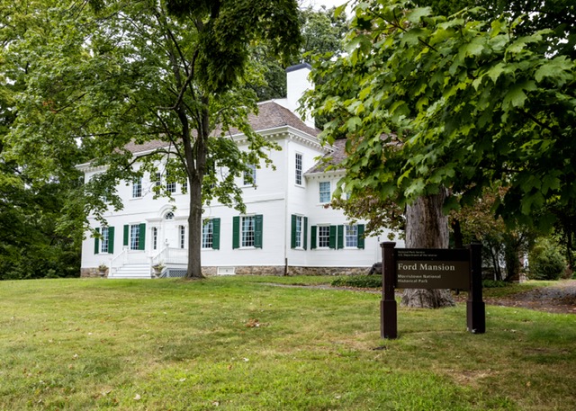 A white two-story colonial house with dark green shutters on the bottom floor stands in the background surrounded by trees. A brown sign stands in the foreground with the title, "Ford Mansion" legible.