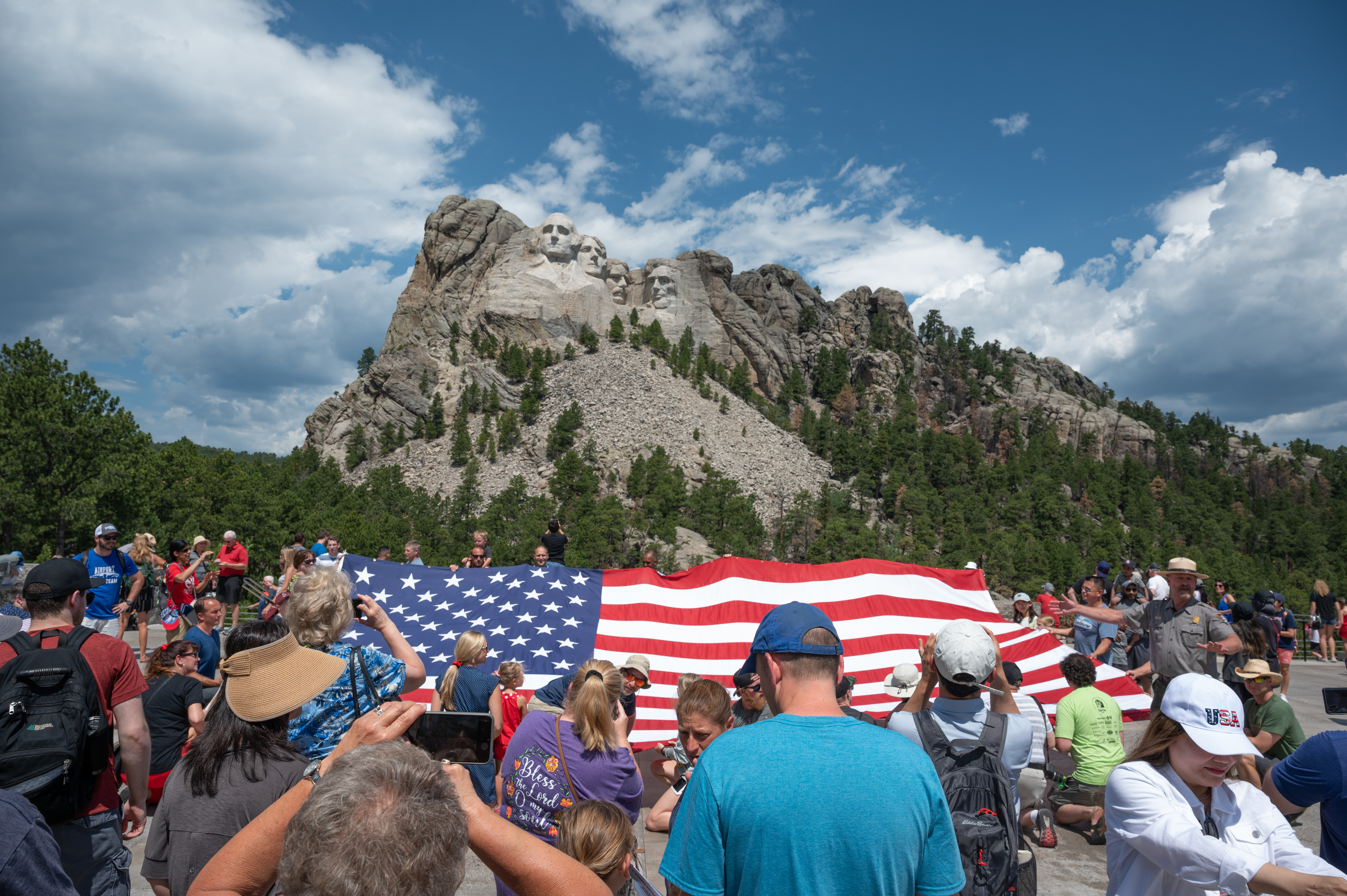Crowd of people standing with a large American flag in front of Mount Rushmore.