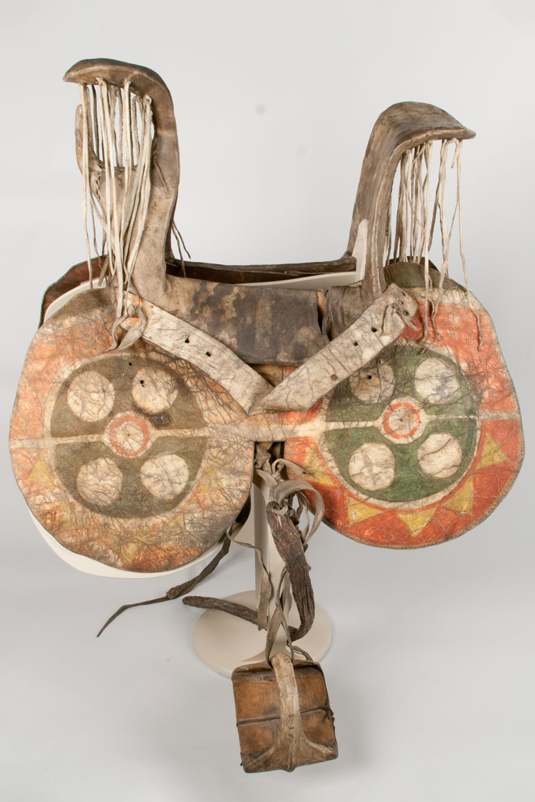 A woman's saddle made from a cottonwood frame with a rawhide covering. The rawhide saddle fenders features geometric designs of green, yellow, and red. Bison hide tie laces secure rawhide inner pieces that form the pemmel and cantle overhangs.