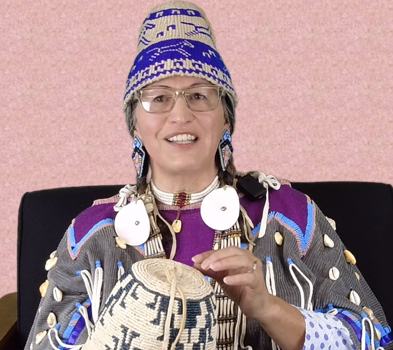 Native American dressed in a decorative shirt. She is wearing a woven hat, earrings, and is holding a basket hat.