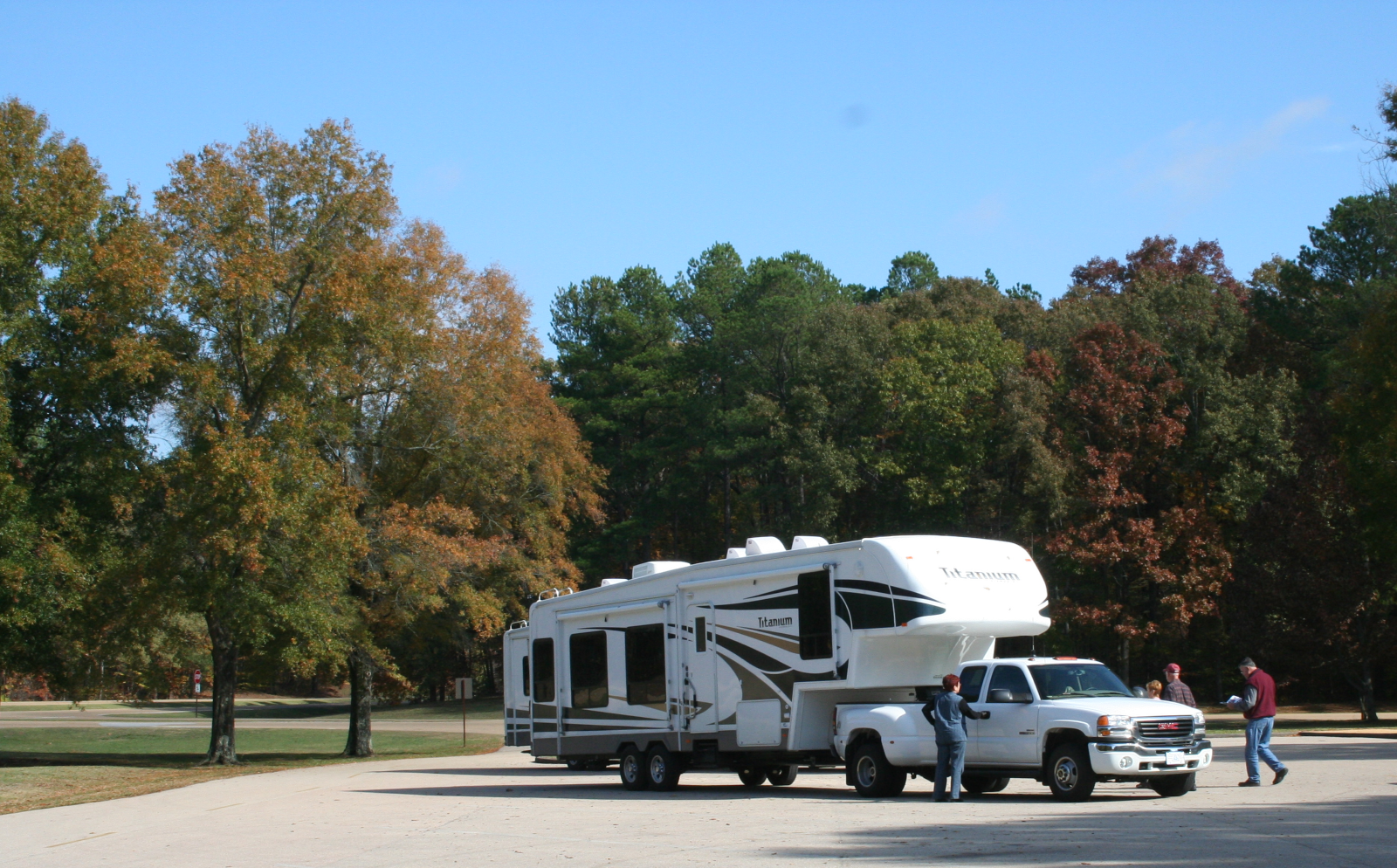 A large white pickup truck towing a large 5th wheel travel trailer is parked in a parking lot. Four people walk toward the vehicle, one looking at a brochure. The lot is surrounded by trees with fall foliage.