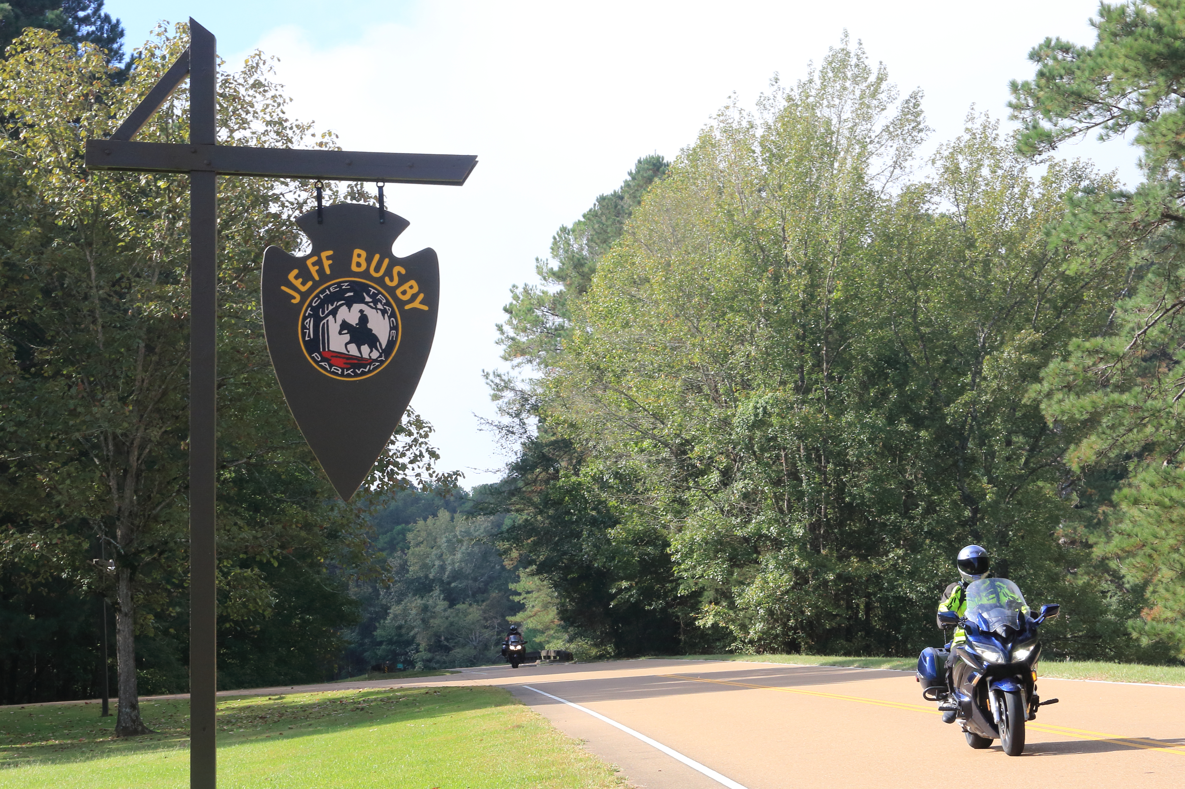 A motorocyclist with helmet and riding suite rides a blue motorcycle on a two lane road. The Jeff Busby brown arrowhead sign is visible in the frame. Another motor cycle is in the distance with its headlights on.