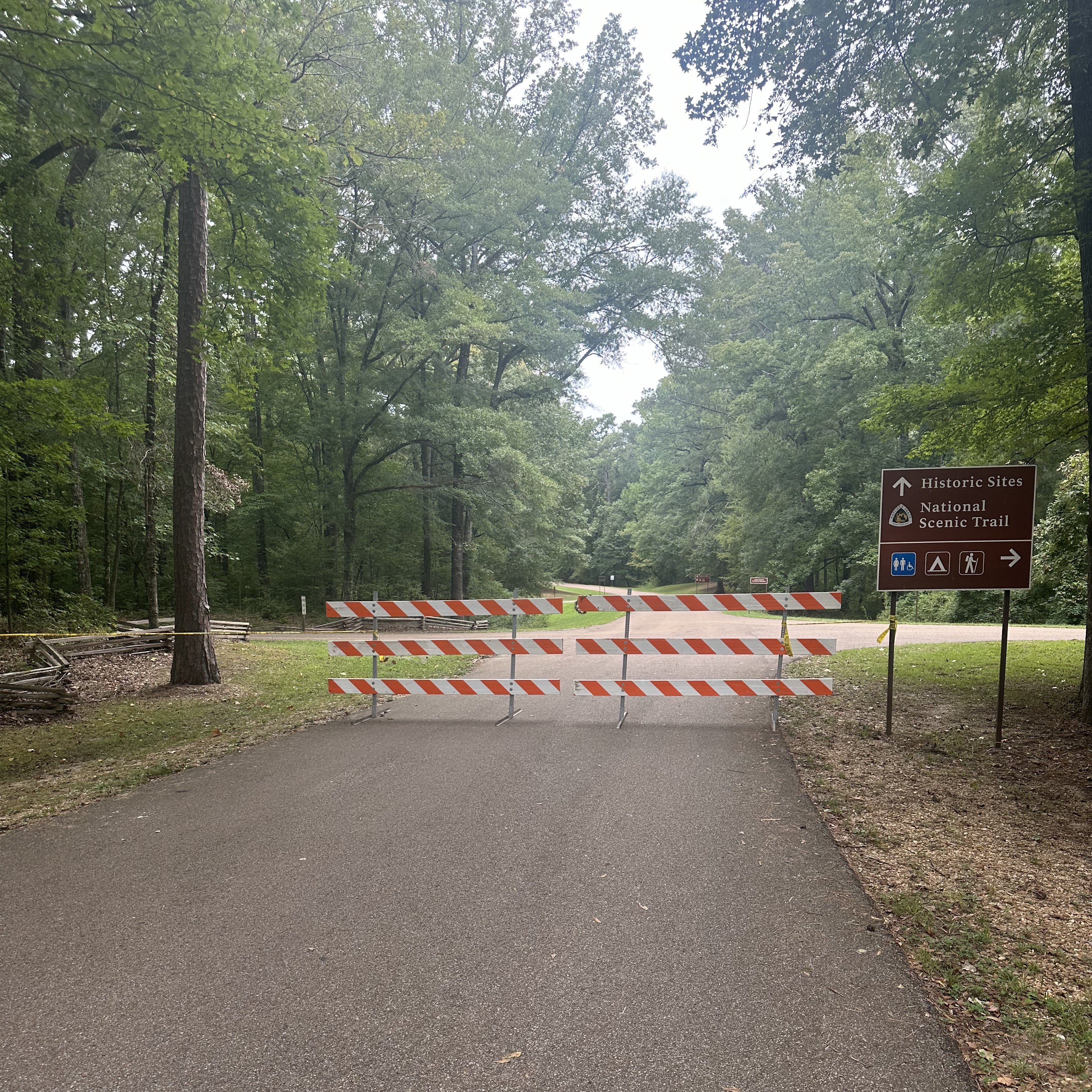 Barricades block the roadway, leading to a wooded area.