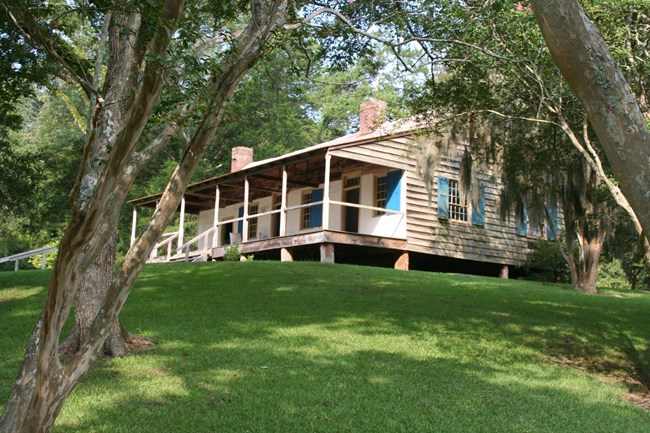 A one story wooden building with blue window shutters stands on wooden blocks atop a grassy hill.