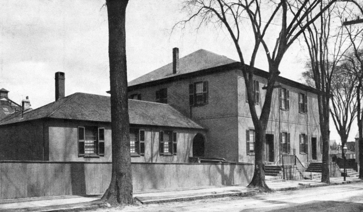Black-and-white photo of meetinghouse, with two entrances, set behind trees.