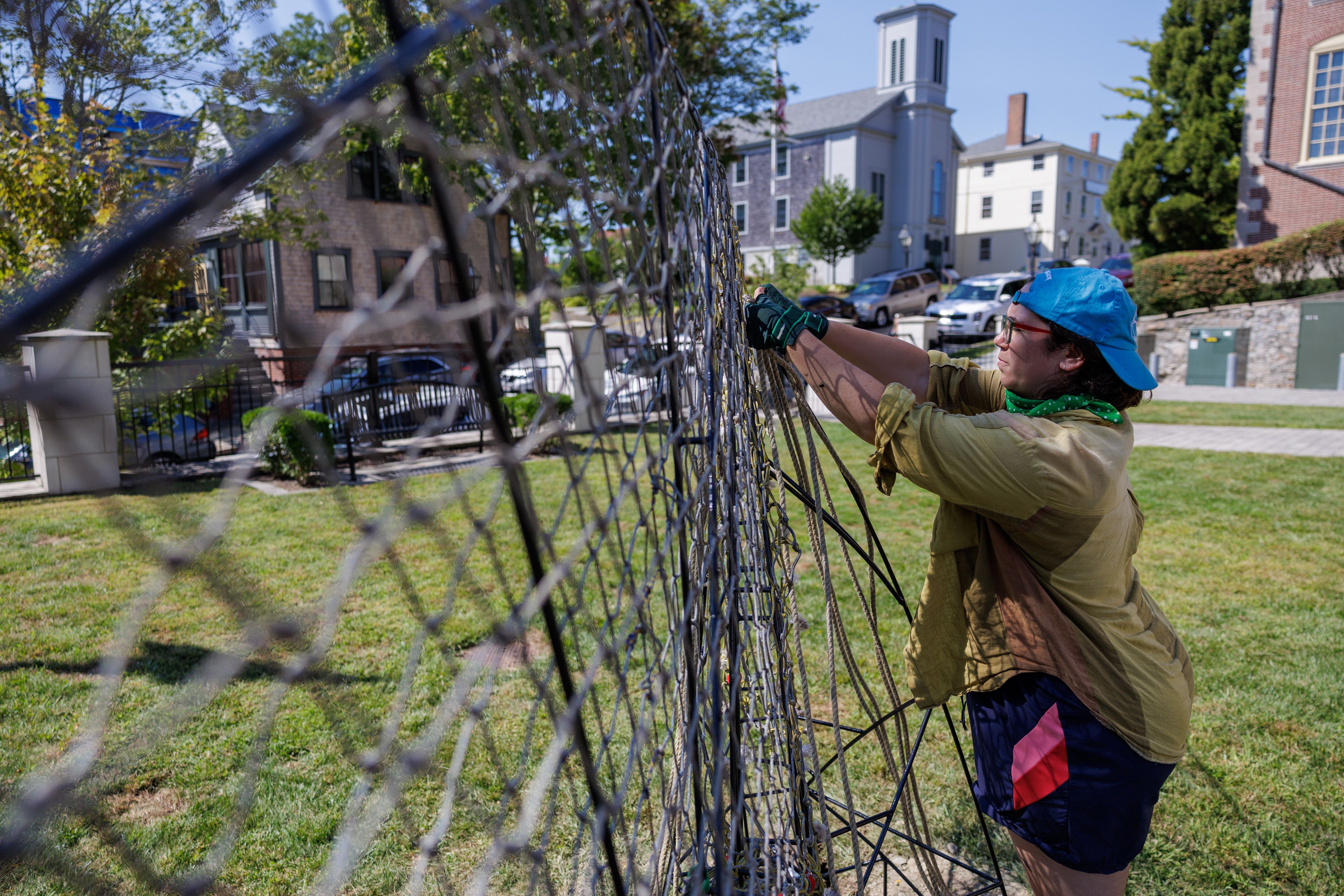 A person in a blue cap and gloves works on a large netted structure in a grassy area near historic buildings, including a church with a white steeple.