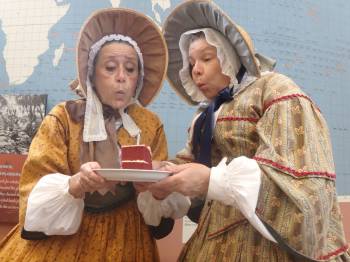 two women in 1850s garb hold a slice of cake and blow out the candle