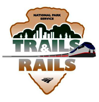 A tan upside down shaped arrowhead with floating text inside that says National Park Service. Underneath, still inside the arrowhead, the words Trails & Rails with a silver, red, and blue Amtrak Train running through the middle.