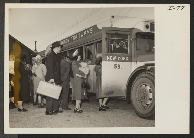 Japanese American families wait in line to board a bus. A man wearing a suit and hat stands behind his wife holding a suitcase. His wife wears a hat and long coat and holds a baby bundled in blankets.