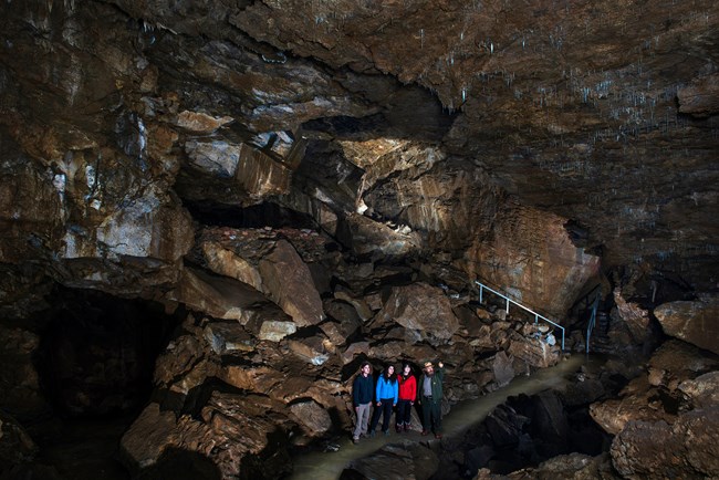 A group of four people stand together inside a large, dimly lit cave. The cave walls and ceiling are jagged and rocky, with visible mineral formations and small stalactites hanging from the ceiling. A narrow walkway and railing are visible in the backgrou