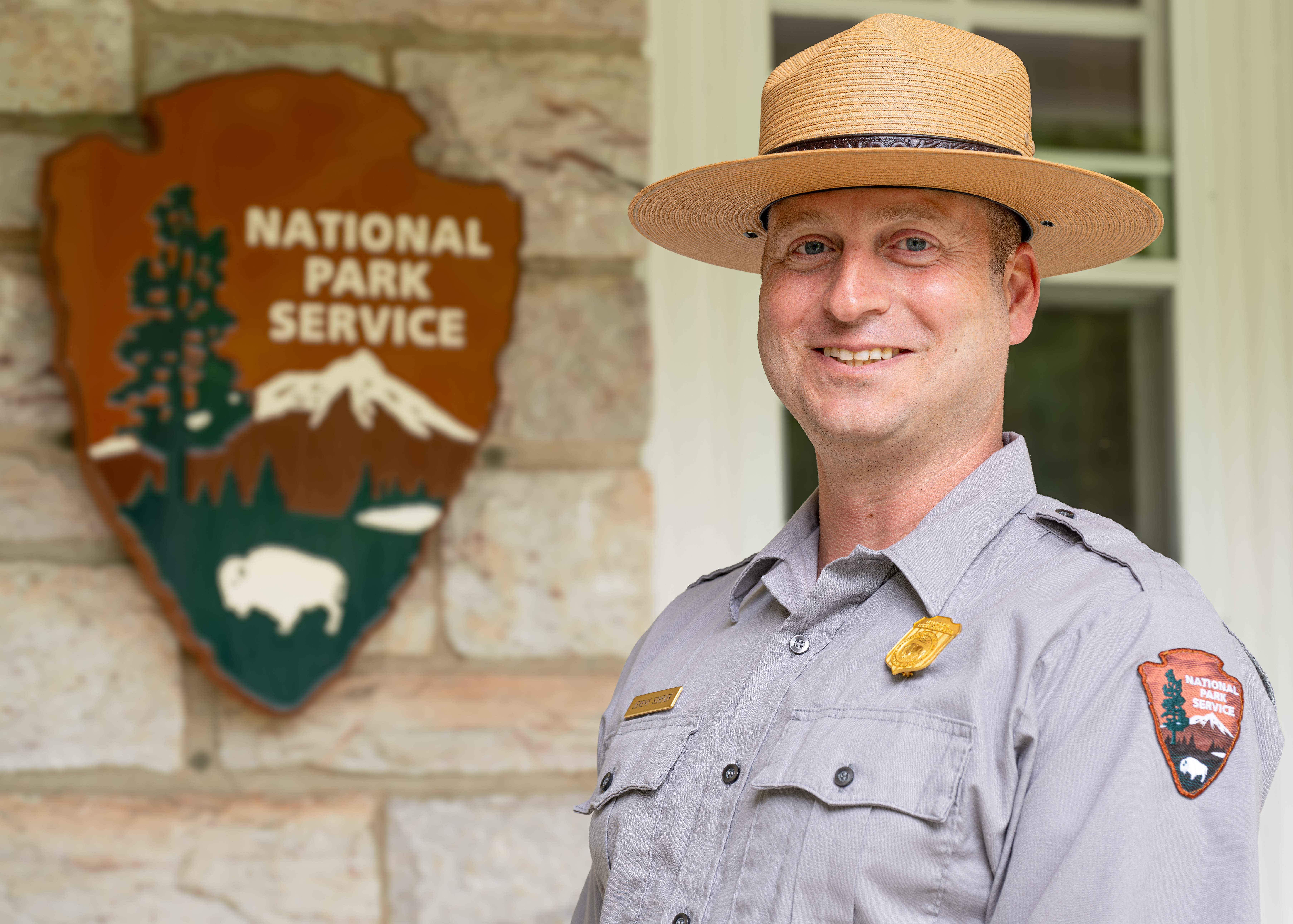 Angled head and shoulders portrait of a white man in a National Park Service uniform standing in front of a stone building with a window and a wooden National Park Service arrowhead behind him.