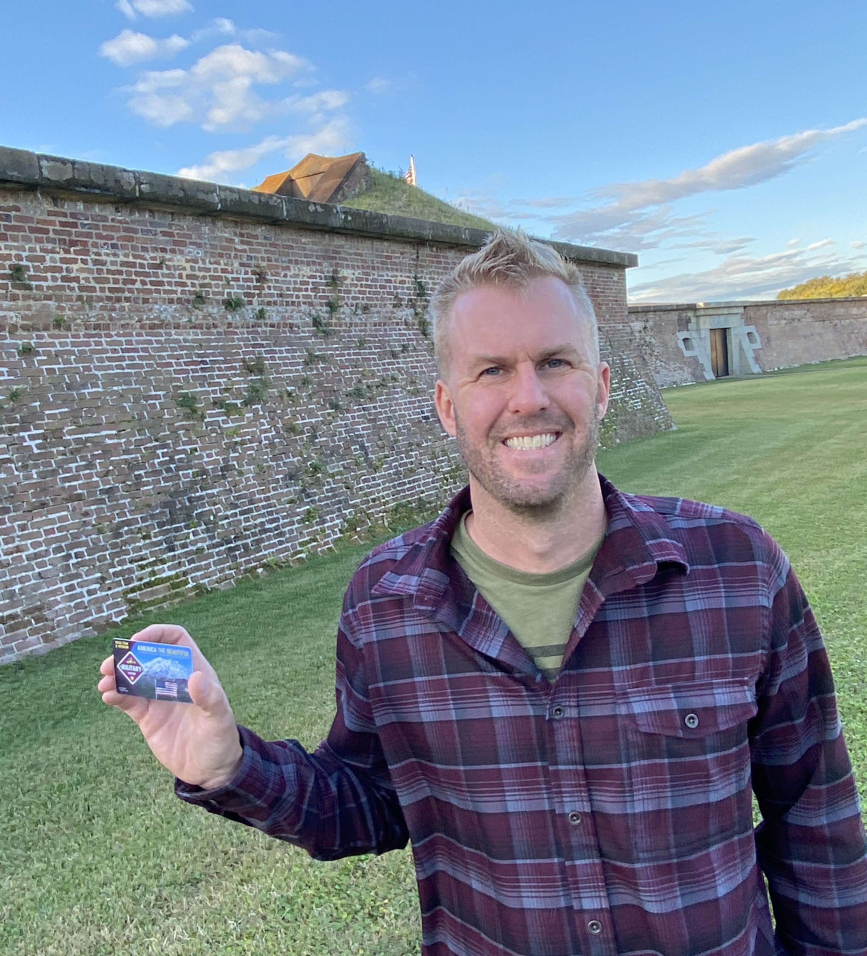 A young man wearing a plaid shirt holds a park pass in his right hand. He is standing on grass in front of the wall of a historic fort.