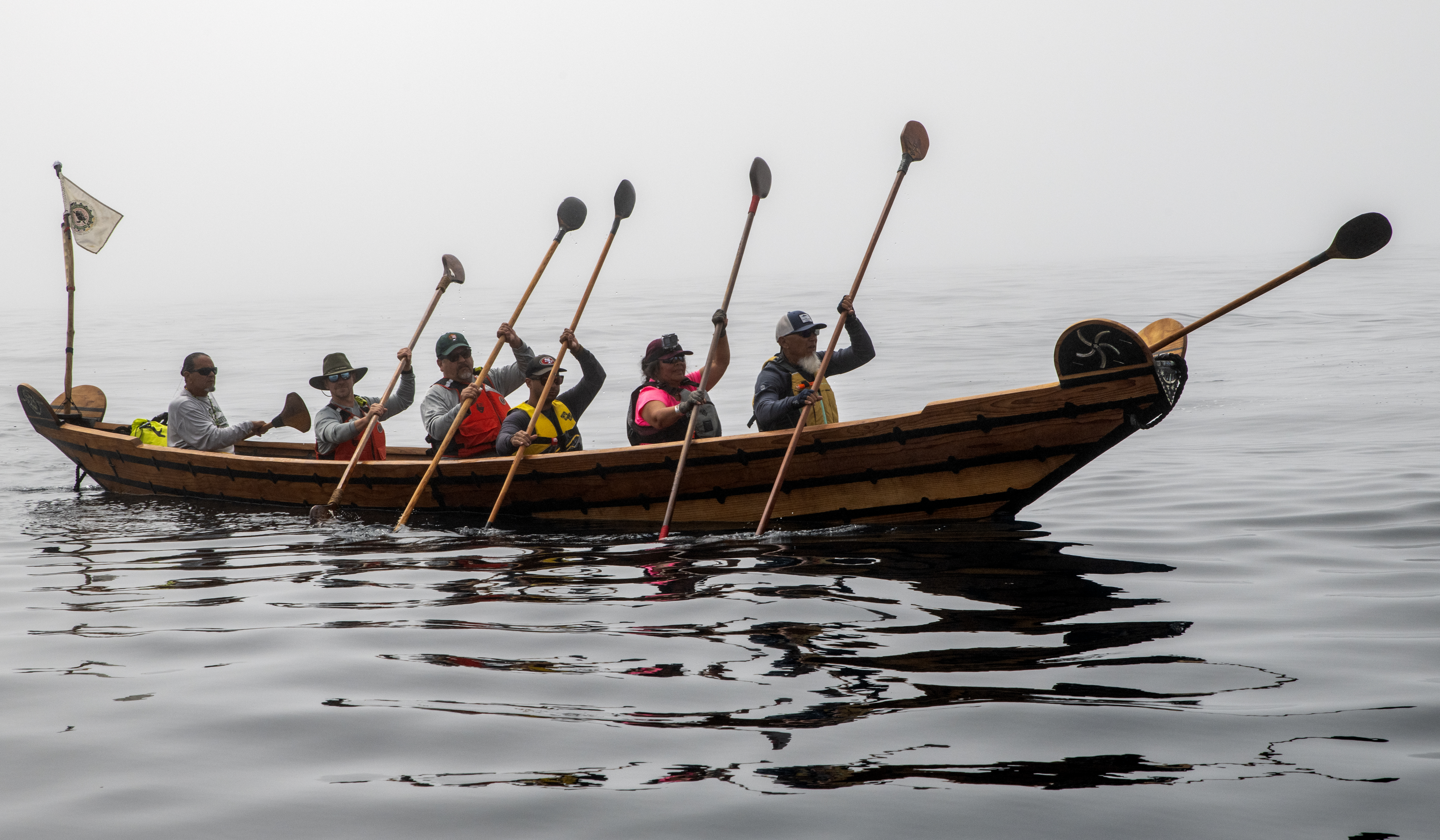 A wooden boat, with a small flag flying at the back, holds 6 people who raise their paddles in unison.