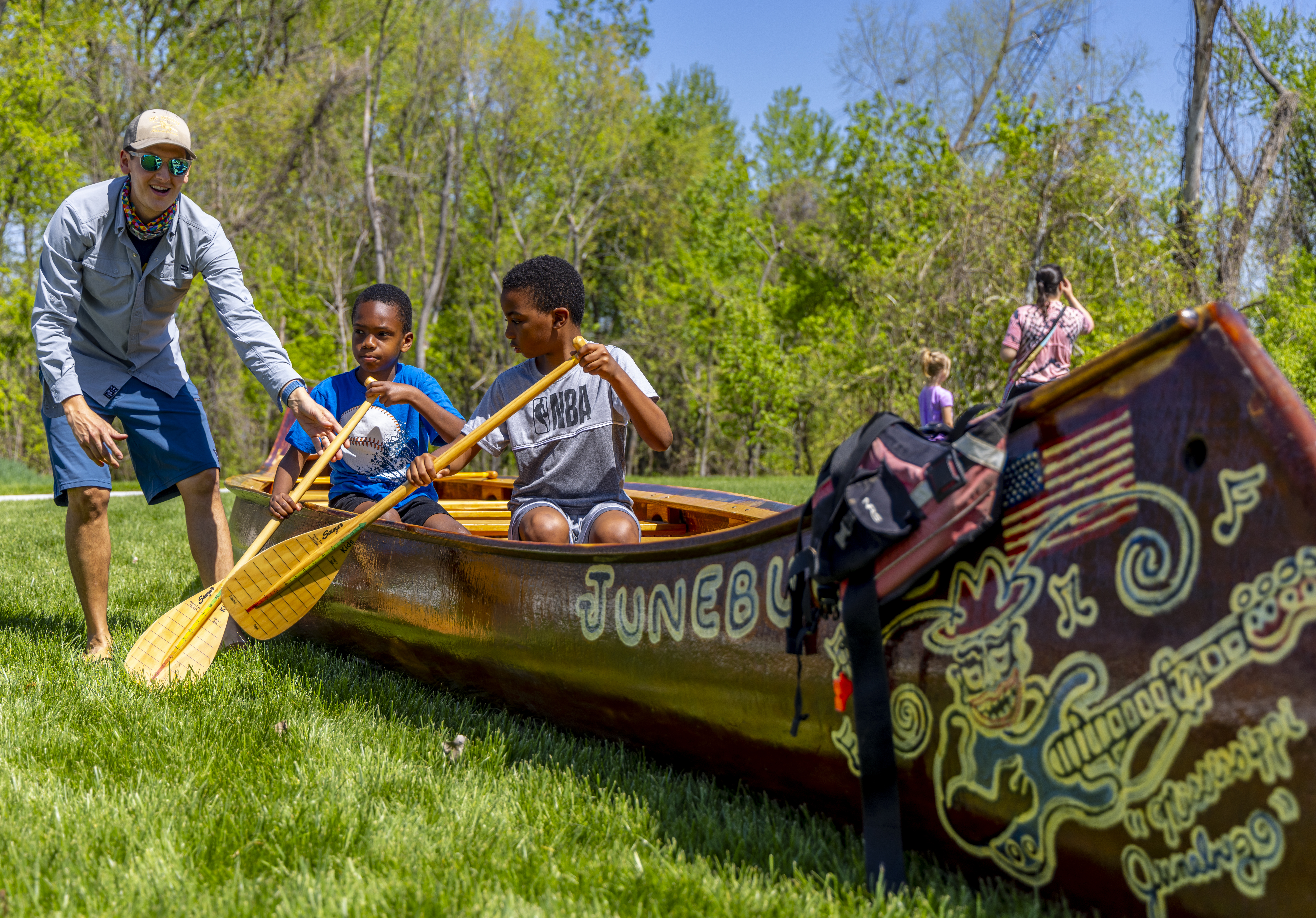 Two boys holding wooden canoe paddles sit in a wooden canoe in a grass field with trees in the background. The canoe has artwork and "JUNEBUG" written on the side of it. A smiling man is standing next to the canoe with one hand on a canoe paddle.