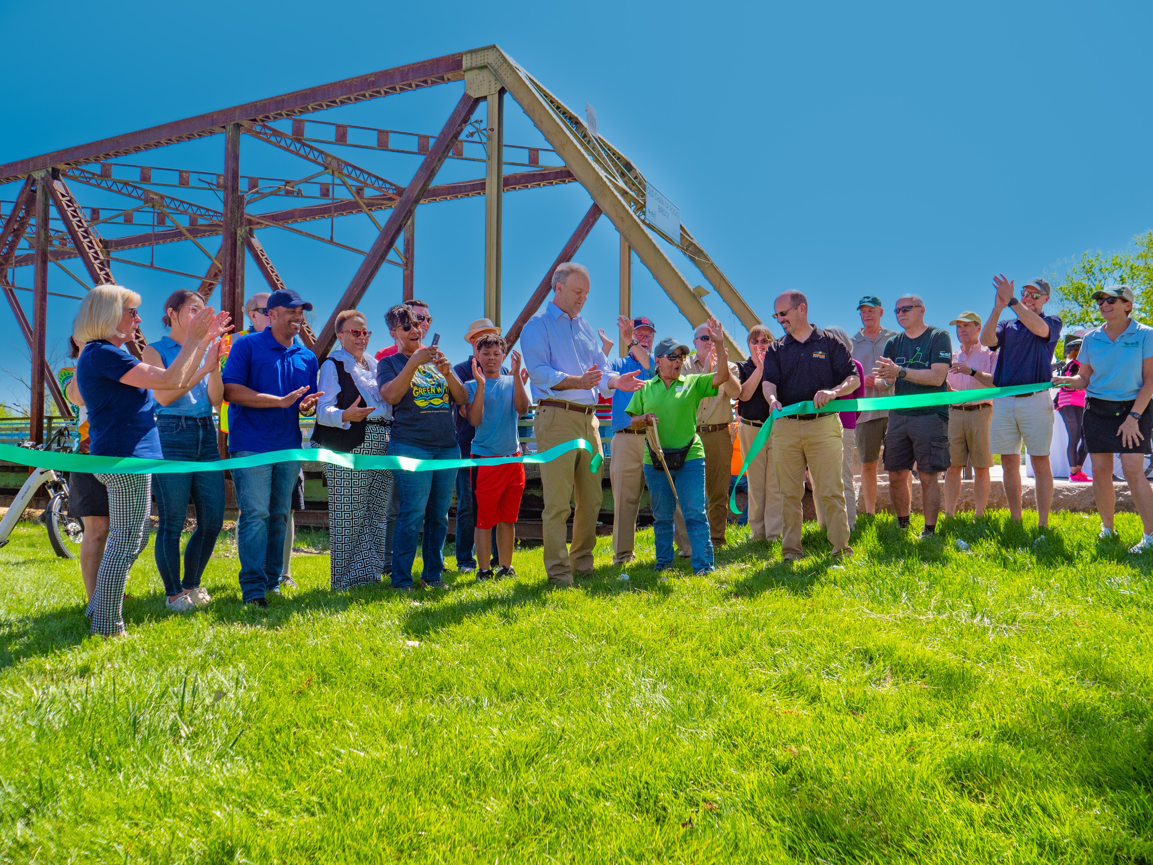 People line up on a bright sunny day to cut a long green ribbon, symbolizing the opening of the park leading to a large brown metal bridge in the foreground