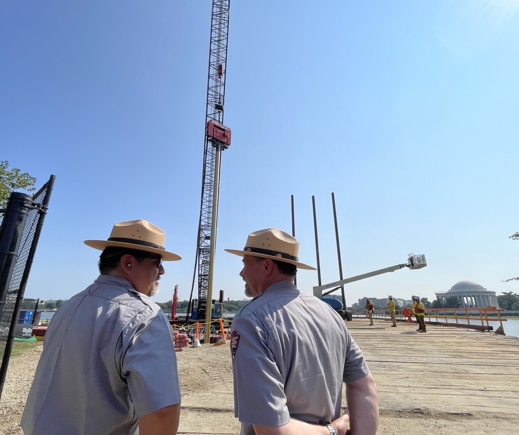 In the foreground, two men wearing park ranger uniforms face one another; in the background, a tall piling reachs up and out of frame.