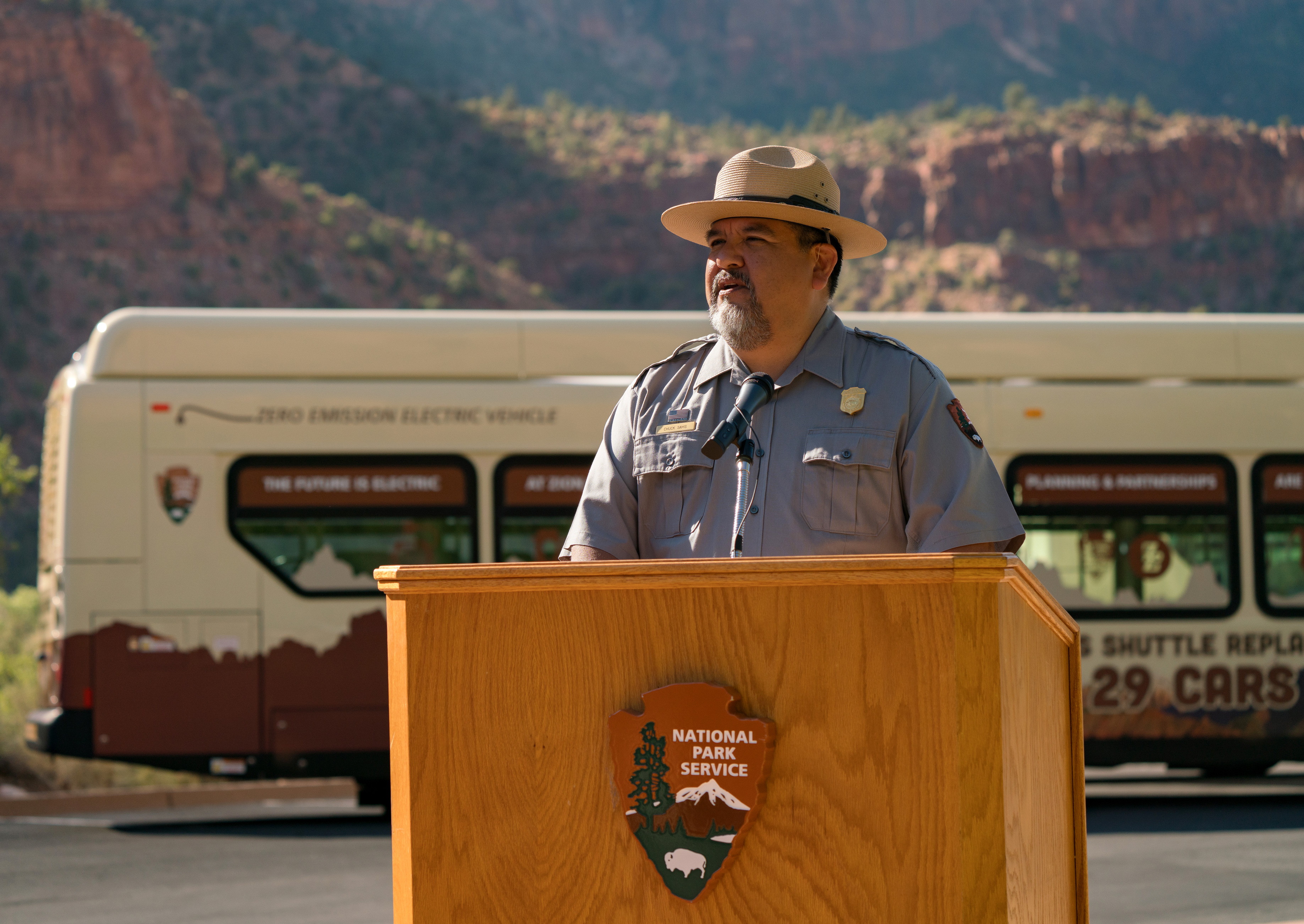 A man in a tan flat-hat, grey uniform shirt with a golden badge and name tag stands behind the microphone of a wooden National Park Service Podium. Behind him, a zero emission electric vehicle is visible.