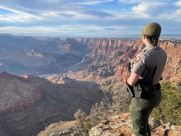 A female law enforcement ranger in uniform stands at a ledge and looks across the Grand Canyon on a partly cloudy day.