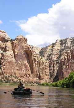 An inflatable raft carrying two people and gear floats down a wide, calm river toward a towering wall of reddish sandstone with white clouds and blue sky overhead.