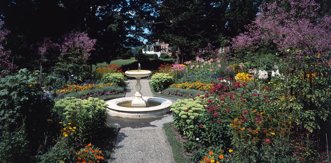 A fountain stands at the axis of a gravel footpath in a formal garden, dense with colorful flowers and shrubs.