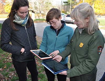 Three women, one in NPS uniform, look at two digital tablets in an autumn landscape.