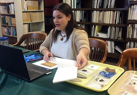 Distance learning session with artifacts Woman seated in front of a laptop computer holding a piece of paper with a tray of artifacts to her left on a table. Shelves of books are behind her.